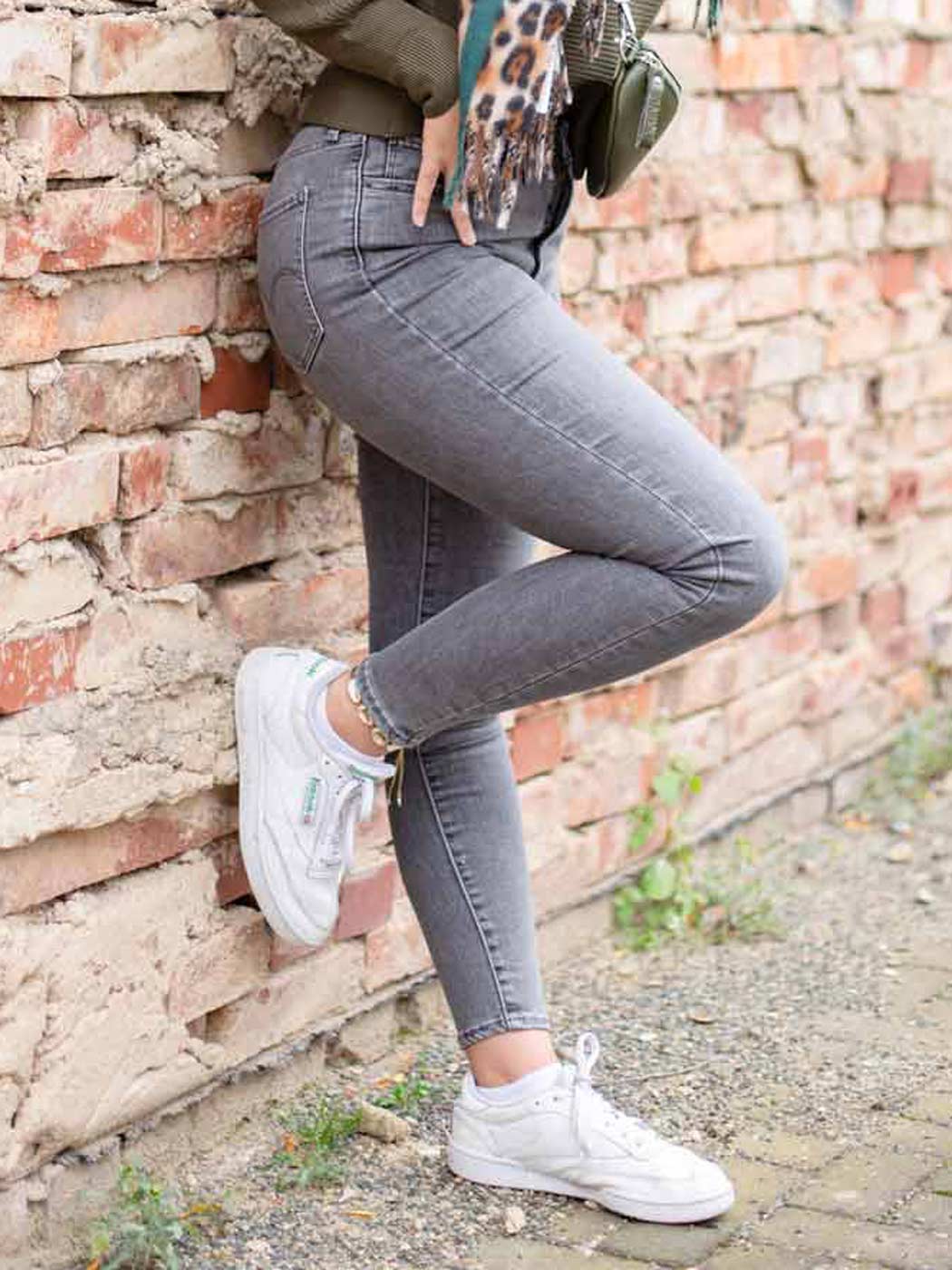 Frau in grauer Skinny Jeans stellt ein Bein angewinkelt an eine Mauer