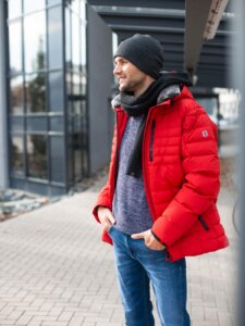 Junger Mann in roter Winterjacke steht vor einem Gebäude mit Glasfront