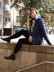 Junge Frau sitzt auf der Mauer und hat eine dunkelblaue Metallicjacke von Wellensteyn an