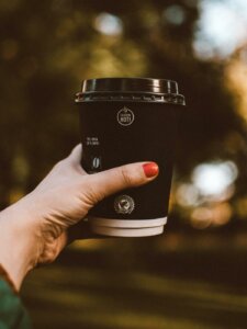 Hand mit lackierten Fingernägeln hält einen Coffee to go-Becher vor einen parkähnlichen Hintergrund