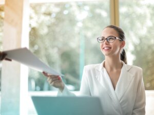 Junge Frau mit Pferdeschwanz und Brille nimmt am Laptop sitzend Papiere entgegen