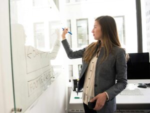 Frau trägt eine helle Bluse und einen grauen Blazer und schreibt mit einem Stift auf eine Tafel