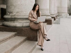 Frau trägt beigen Blazer und eine beige Anzughose, sie sitzt auf einem Stein und sieht auf ihren Laptop