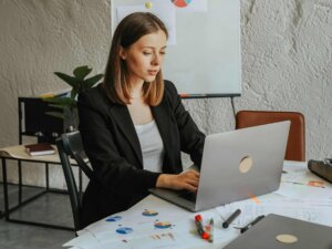 Frau sitzt an einem Schreibtisch und schreibt an ihrem Laptop; sie trägt ein weißes Top und einen schwarzen Blazer