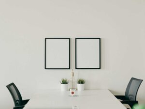 Ein weißer Tisch, an dem zwei schwarze Stühle stehen und wenig Dekoration, wie schwarze Bilderrahmen mit weißem Hintergrund