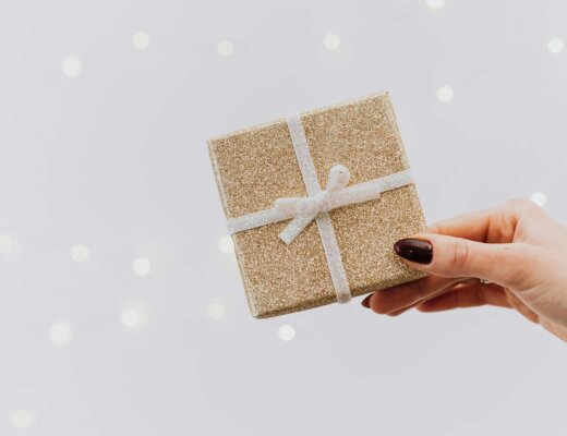 Vor einem grauen Hintergrund wird ein in goldenes Papier eingeschlagenes viereckiges Geschenk gehalten.