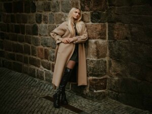 Eine Frau trägt schwarze Overknee-Stiefel zu einem schwarzen Kleid und einem langen, beigen Mantel.