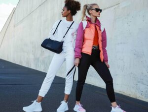 Zwei Frauen stehen mit dem Rücken zueinander. Eine trägt ein komplett weißes, sportliches Outfit und die andere eine schwarze Leggings und eine neonfarbene Jacke.
