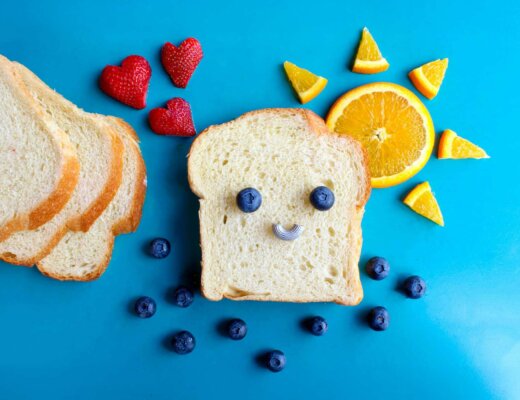 Ein blauer Hintergrund. Zu sehen ist eine Toastscheibe, auf der mit Blaubeeren ein lächelndes Gesicht gelegt wurde. Daneben ist eine Sonne aus einer Orange geschnitten worden und Erdbeeren in Herzform.