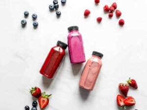 Drei rosafarbene und rote Smoothies liegen in Glasflaschen abgefüllt neben verschiedenen Beeren.