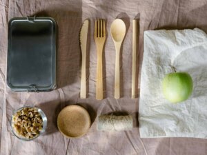 Eine metallene Brotdose, Holzbesteckt, ein Apfel und ein Tuch liegen nebeneinander.