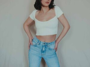 Eine Frau steht vor einer grauen Wand und trägt eine hellblaue Jeans und ein weißes cropped Oberteil mit einem großen Ausschnitt.