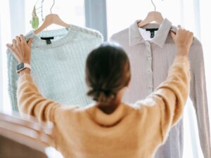 Eine Frau steht vor einem Fenster und hält zwei Oberteile in die Luft und guckt zwischen diesen hin und her, um zu entscheiden, welches sie tragen soll.