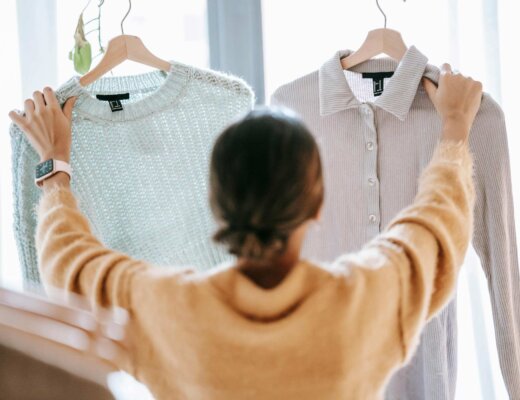 Eine Frau steht vor einem Fenster und hält zwei Oberteile in die Luft und guckt zwischen diesen hin und her, um zu entscheiden, welches sie tragen soll.