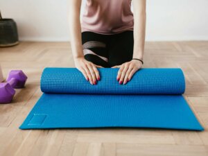 Eine Frau in Sportklamotten sitzt auf dem Boden und rollt eine blaue Sportmatte aus.