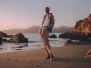 Ein großer schlanker Mann steht am Strand und posiert mit seinen Händen in den Hosentaschen.