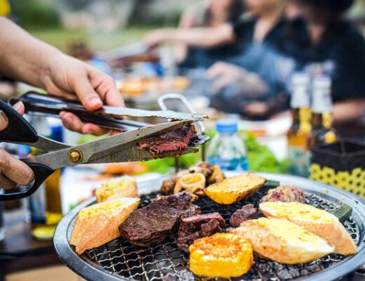 Auf einem Grill liegen Fleischstücke und Baguettescheiben, im Hintergrund sieht man verschwommen Leute bei einem Grillfest.