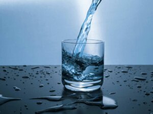 In ein Glas wird Wasser eingeschüttet, drumherum sind überall Wassertropfen und kleine Pfützen.
