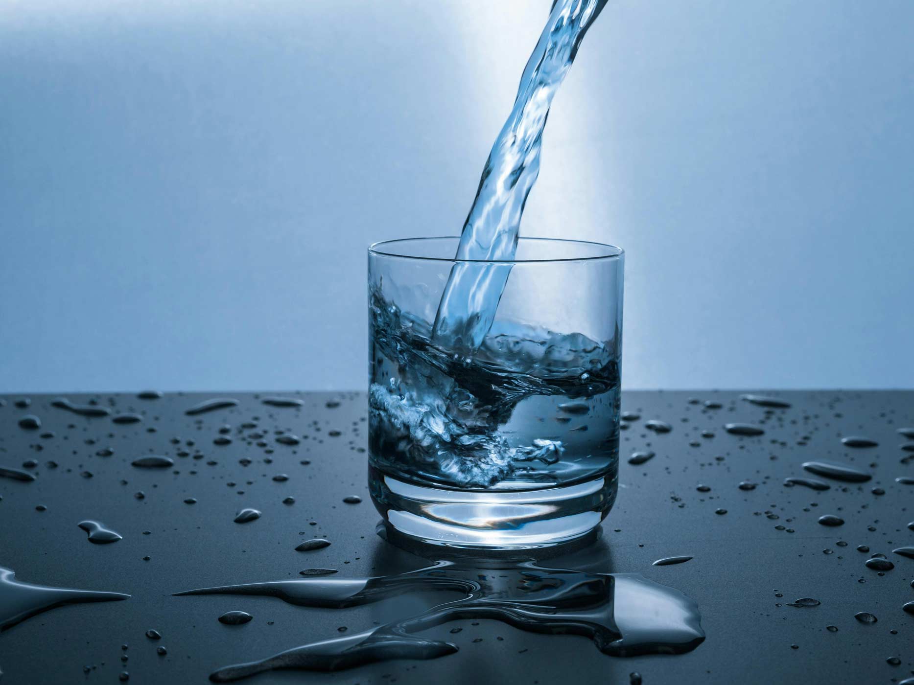 In ein Glas wird Wasser eingeschüttet, drumherum sind überall Wassertropfen und kleine Pfützen.