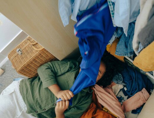 Eine Frau liegt vor ihrem Kleiderschrank und auf ihr drauf liegen viele Klamotten.