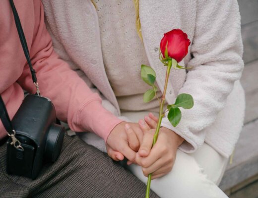 Man sieht die verschlungenen Hände von einem Mann und einer Frau, die zusammen auf einer Holzbank sitzen. Die Frau hält eine rote Rose in der Hand.