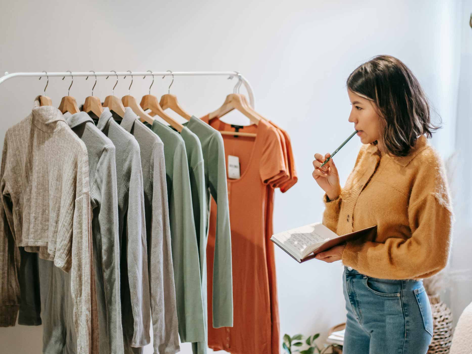 Eine Frau steht vor einer Kleiderstange mit Klamotten, sie hält ein Buch in der Hand und kaut auf einem Stift.