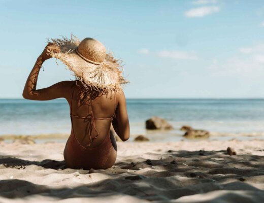 Eine Frau im Bikini sitzt am Strand. Die Sonne scheint und sie hält den Hut, den sie trägt, fest.