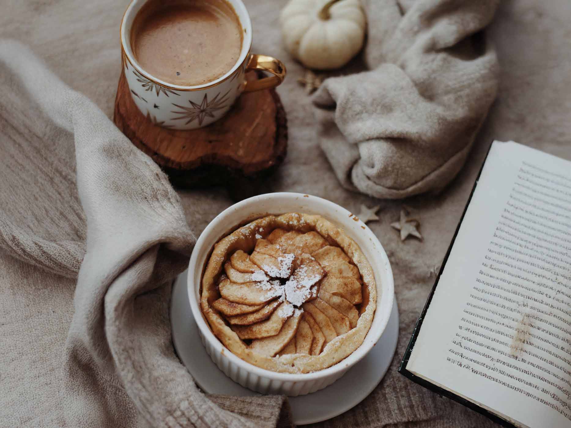 Herbst Gebäck Rezepte Ein Apfelkuchen steht mit einer Tasse Kakao auf einem Tisch. Daneben liegen ein Pullover, ein Buch und ein kleiner Kürbis.