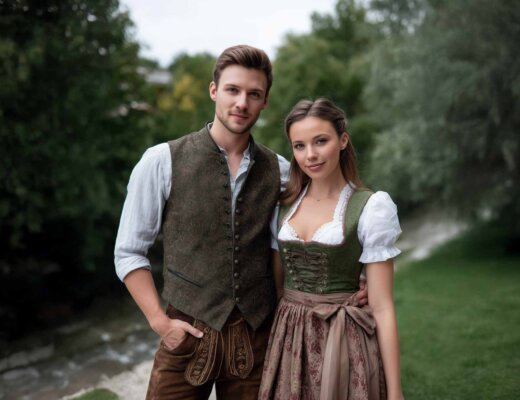 Ein Mann und eine Frau stehen nebeneinander. Der Mann trägt ein traditionelles Outfit mit Lederhose. Die Frau trägt ein Dirndl. Beide Lächeln glücklich.