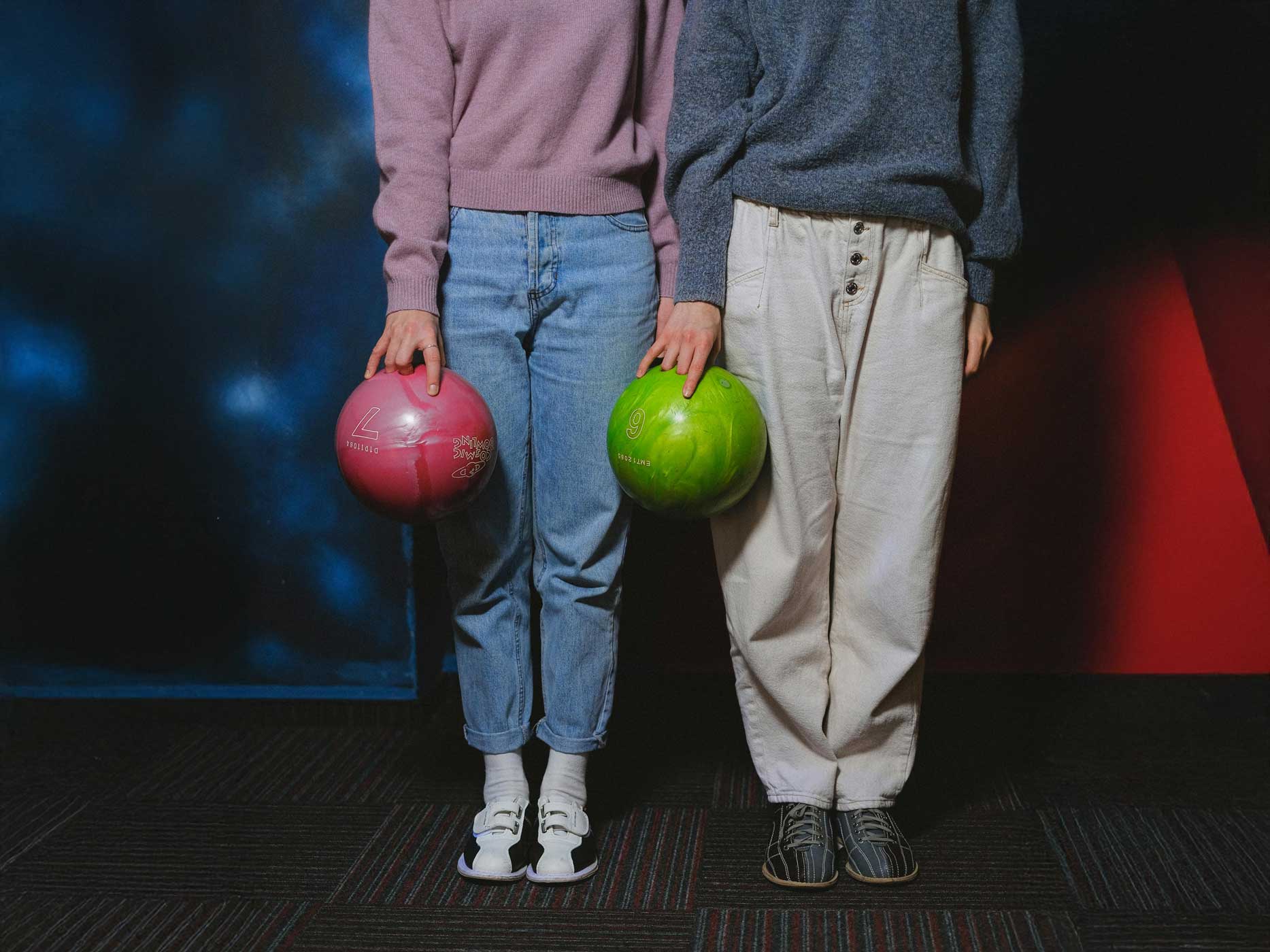 Passendes Outfit Bowling Zwei Frauen stehen nebeneinander und halten jeweils eine Bowlingkugel in der Hand, die eine trägt eine blaue Mom-Jeans und einen lockeren rosanen Pullover und die andere eine beige Stoffhose und einen dunklen grau-blauen Pullover.