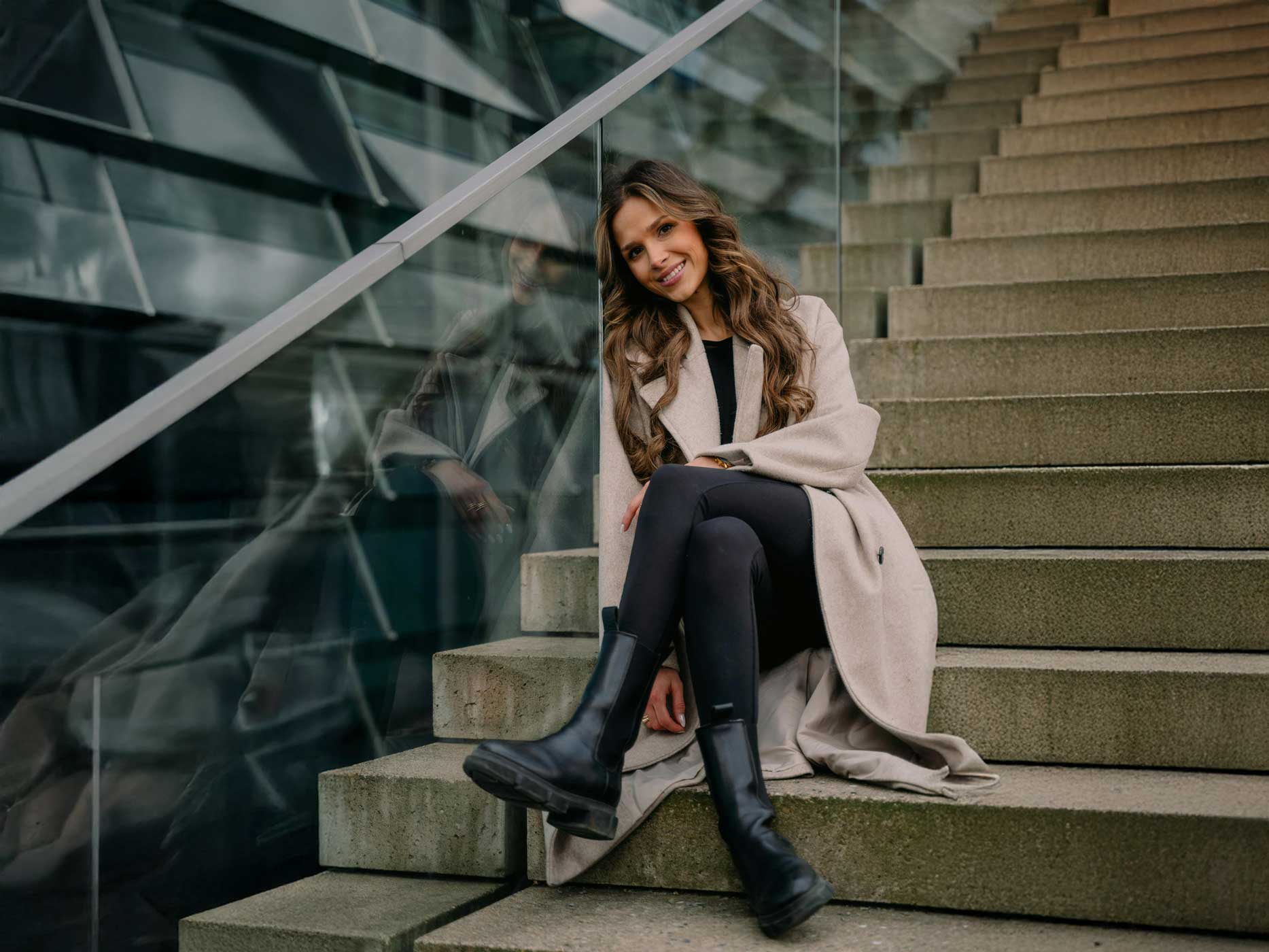 Passendes Outfit saisonal Eine Frau sitzt auf einer Treppe, sie trägt eine schwarze Leggings, ein schwarzes Oberteil, schwarze Stiefel und einen beigen/braunen Mantel.