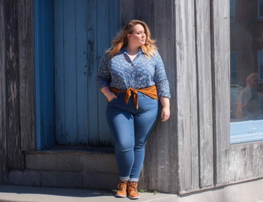 Eine Frau lehnt an einem Gebäude. Sie trägt eine blaue Skinny Jeans und hat ein Tuch als Gürtel umgebunden.