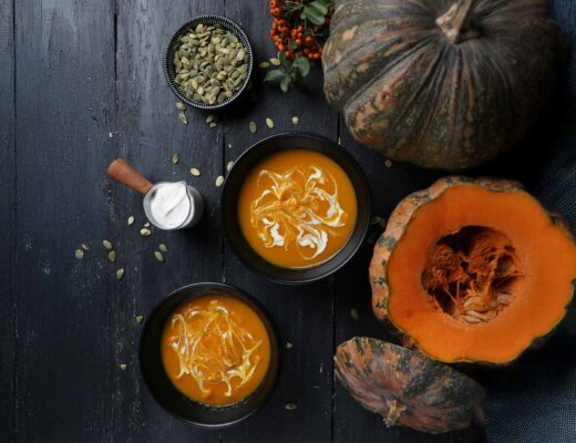 Auf einem Tisch stehen zwei Schalen mit Kürbissuppe und eine mit Kürbiskernen. Außerdem liegen daneben noch ganze Kürbisse.