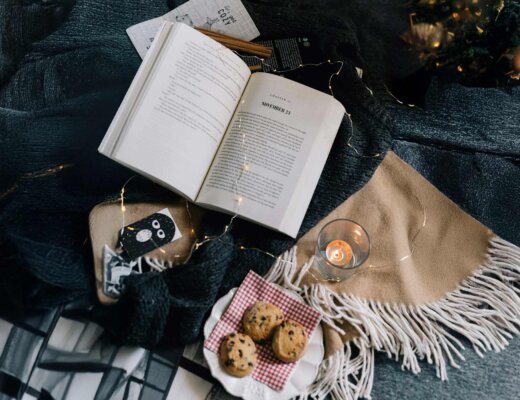 Auf einer Tischdecke liegen ein aufgeschlagenes Buch, ein Teller mit Keksen, eine Lichterkette und Kerzen.