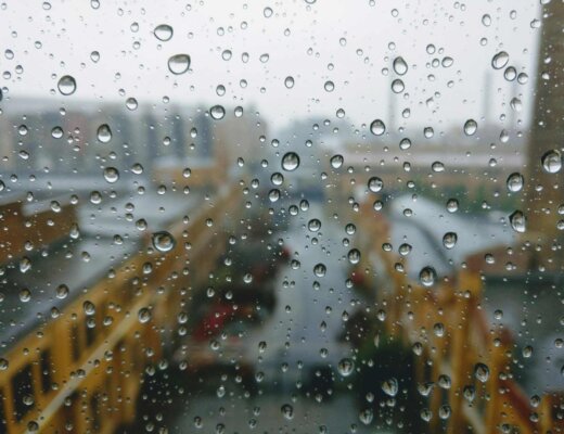 An einem Fenster mit Aussicht auf eine Innenstadt prallen Regentropfen ab.