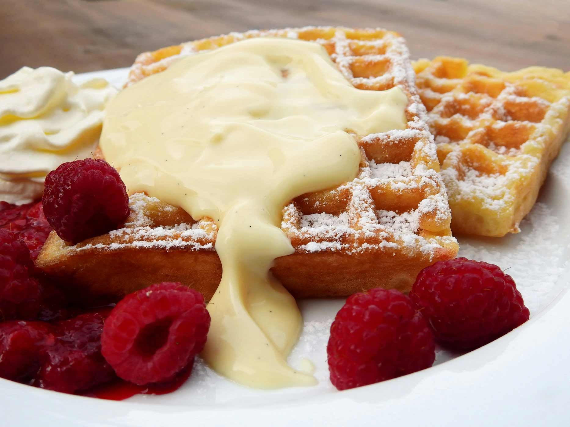 Geburtstag Kuchen backen Vanillecreme zerläuft über mit Puderzucker bestäubtes Waffelherz, neben dem Himbeeren liegen.