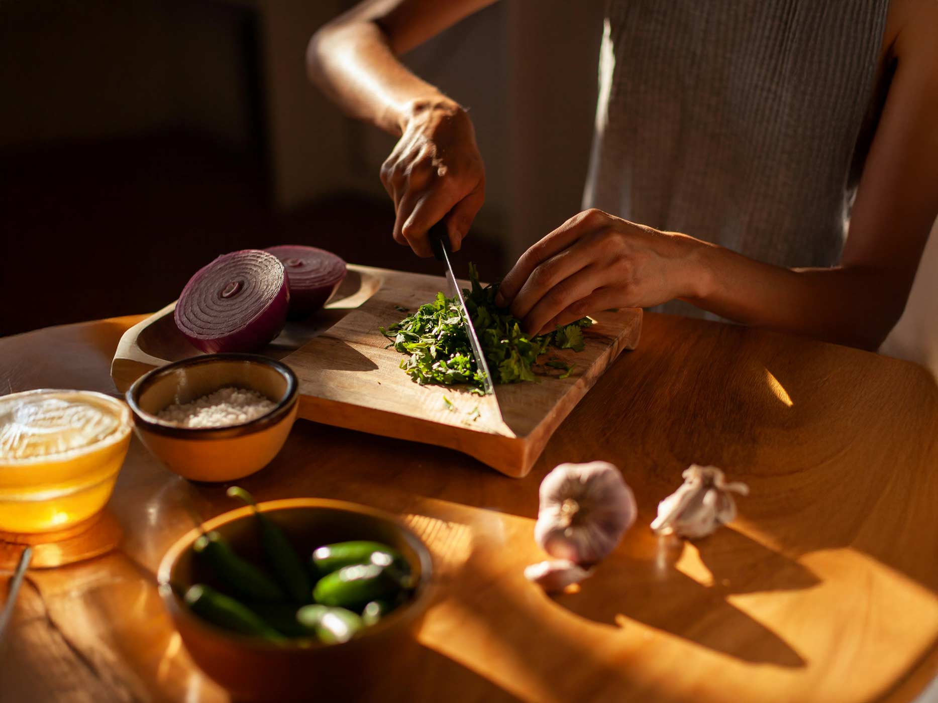 Life Hacks Küche Eine Frau sitzt an einem Holztisch, auf den die Sonne scheint, und schneidet frische Petersilie. Auf dem Tisch stehen weitere Lebensmittel wie Zwiebeln.
