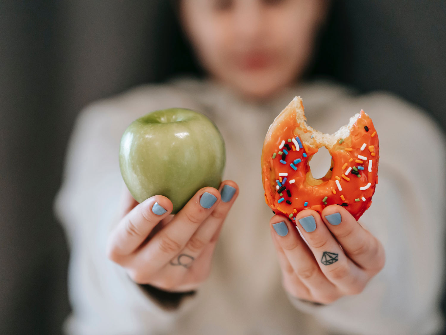 Frau hält einen grünen Apfel und einen bunten Donut in die Kamera