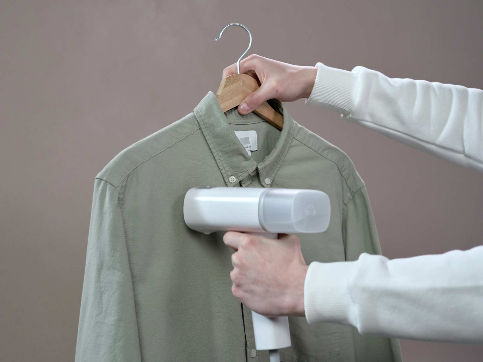 Ein Mann hält ein Hemd auf einem Bügel in der einen Hand und in der anderen einen tragbaren Steamer, mit dem er das Hemd glättet