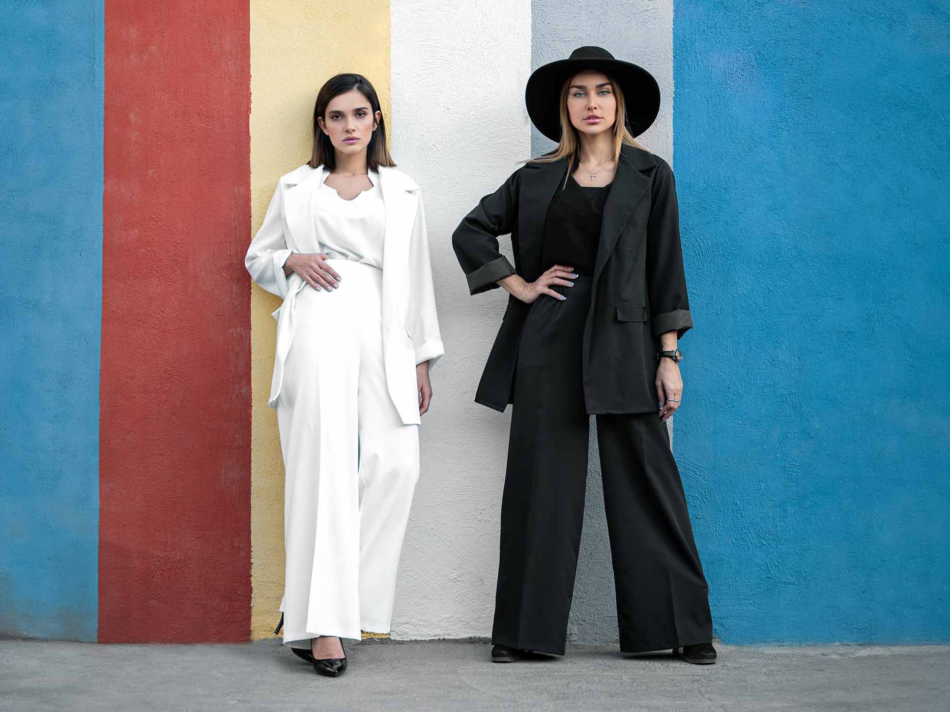 Eine Frau in einem komplett weißen Outfit und eine Frau in einem komplett schwarzen Outfit stehen vor einer Wand