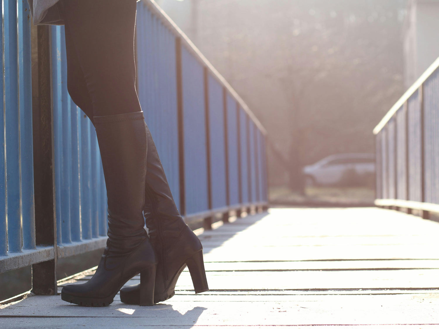 Frau steht bei Sonnenschein auf Holzbrücke und trägt dabei eine schwarze Hose und schwarze Overknees mit Absatz