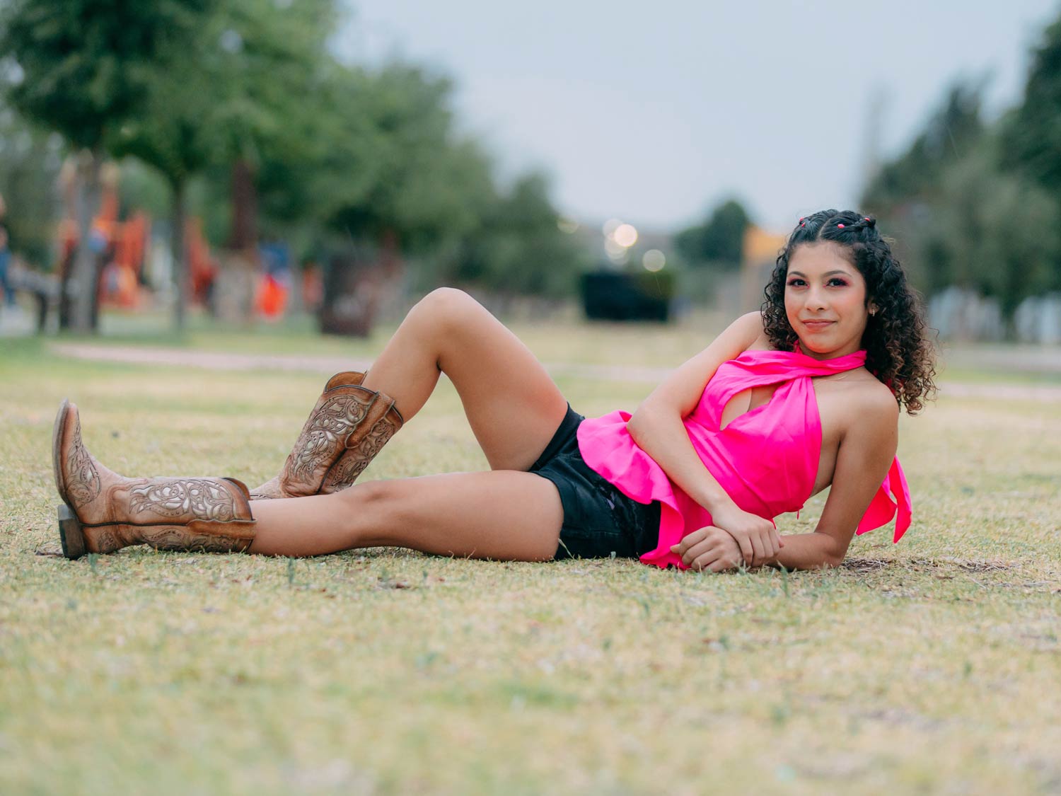 Frau liegt auf dem Arm aufgestützt in pinkem Oberteil und schwarzer, kurzer Hose mit braunen Cowboystiefeln auf dem Rasen