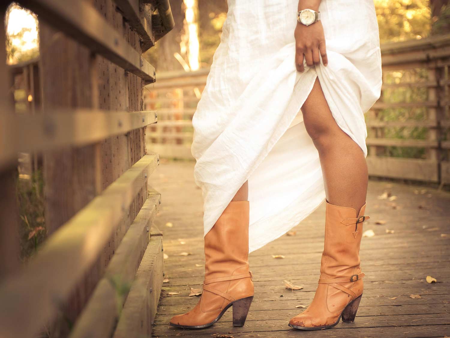 Frau steht bei Sonnenschein auf einer Holzbrücke und trägt ein leicht fallendes, weißes Kleid, hebt dieses an und zeigt dadurch ihre braunen Boots