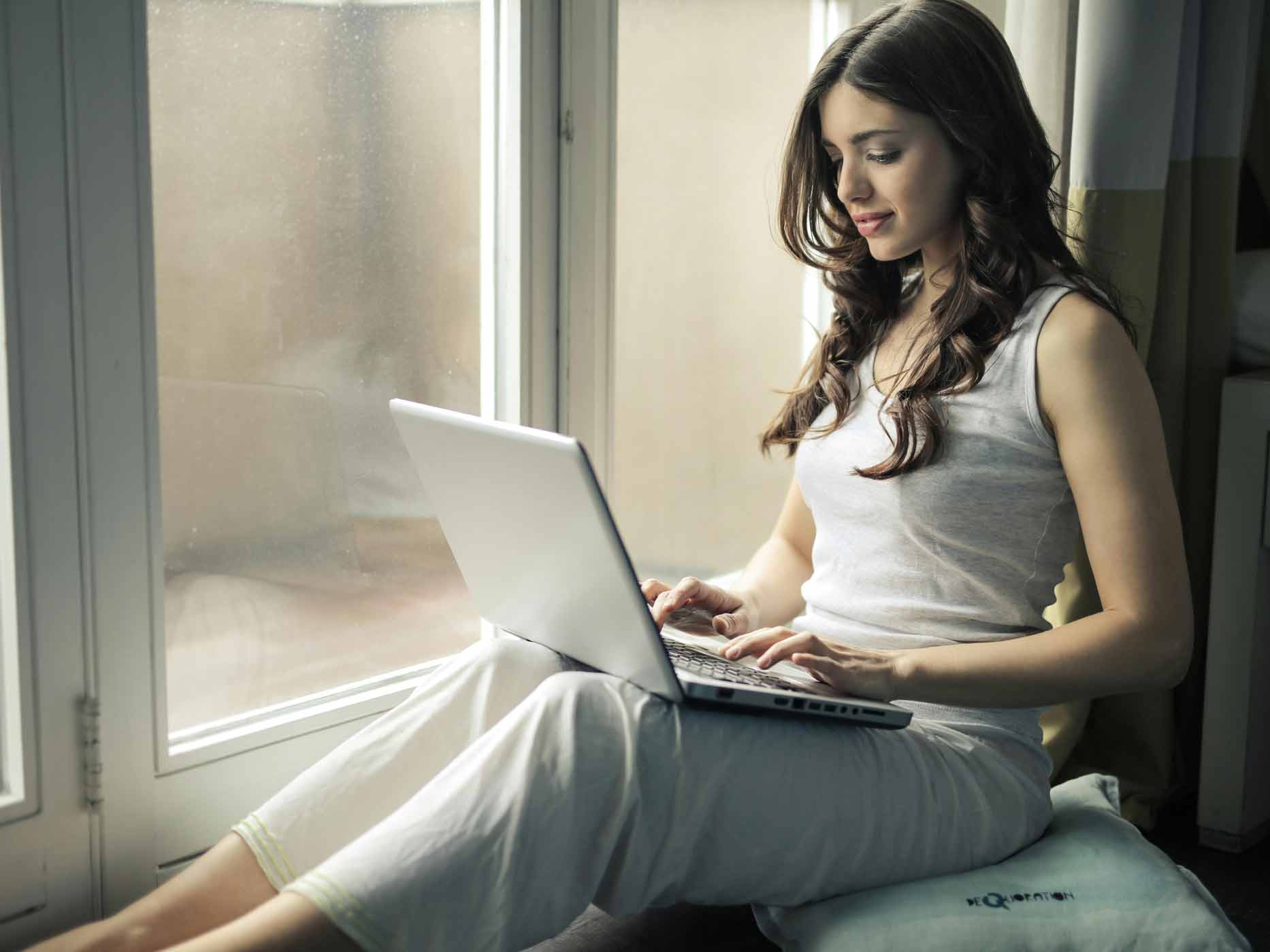 Frau sitzt mit einem Laptop auf den Beinen in einer Fensternische