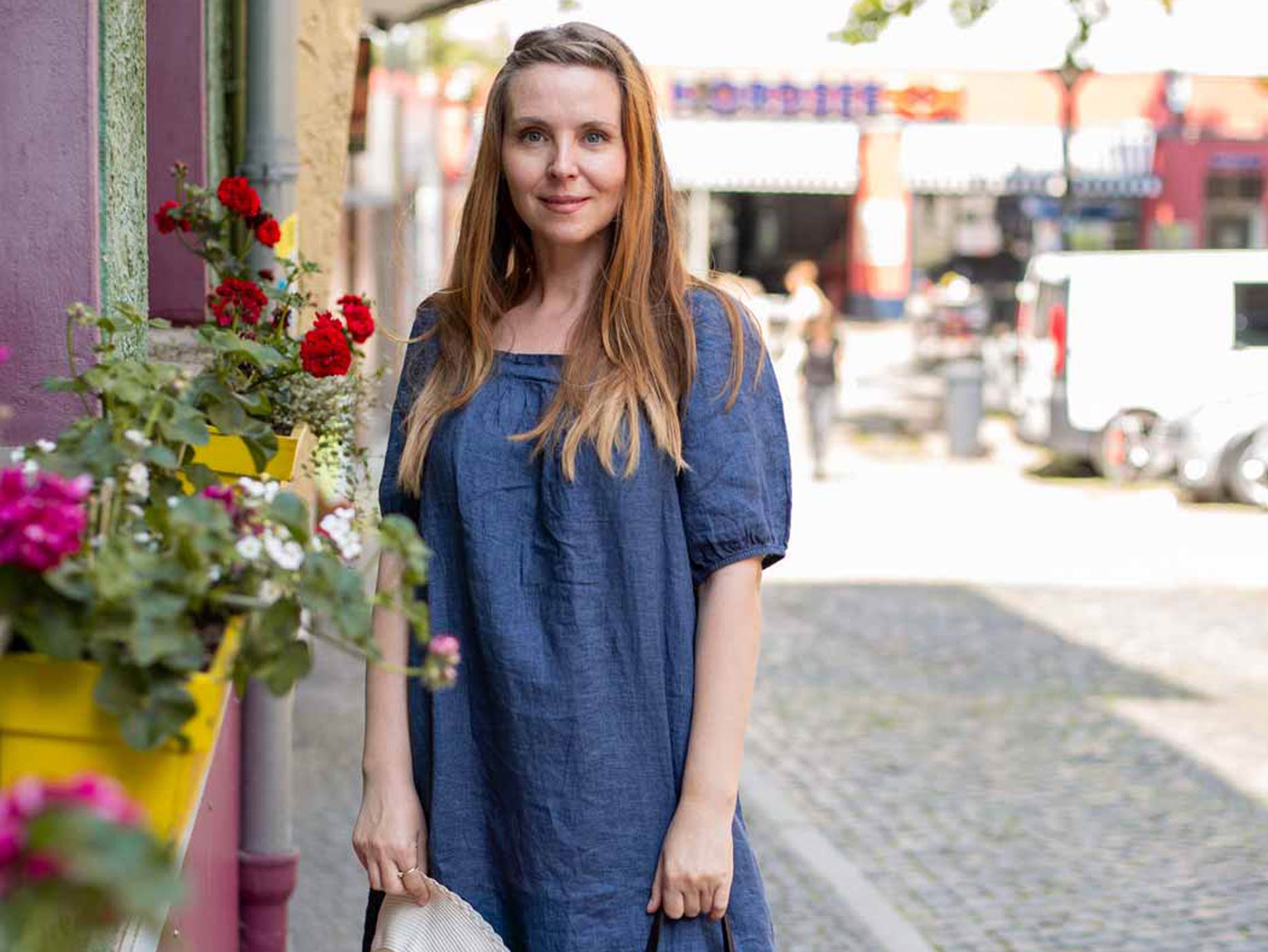 Eine Frau trägt ein leichtes, dunkelblaues Sommerkleid und steht in der Stadt vor Blumen