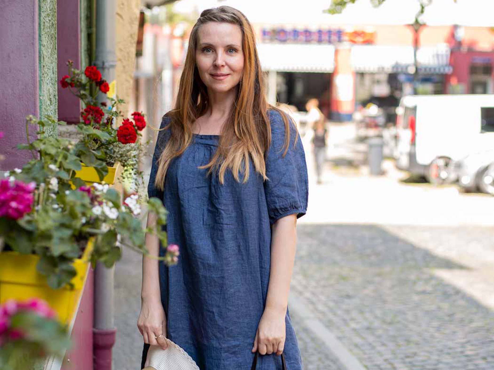 Frau trägt leichtes, dunkelblaues Sommerkleid und steht vor Blumen in der Stadt