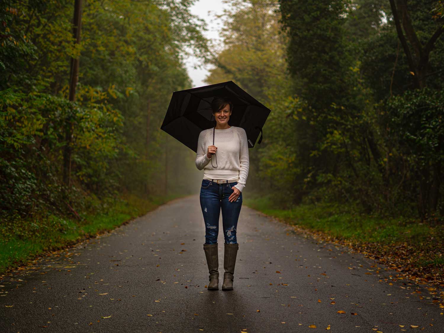 Frau mit blauer Jeans und weißem Oberteil steht mit Stiefeln und einem Regenschirm in der Hand auf einem Weg vor dem Wald.