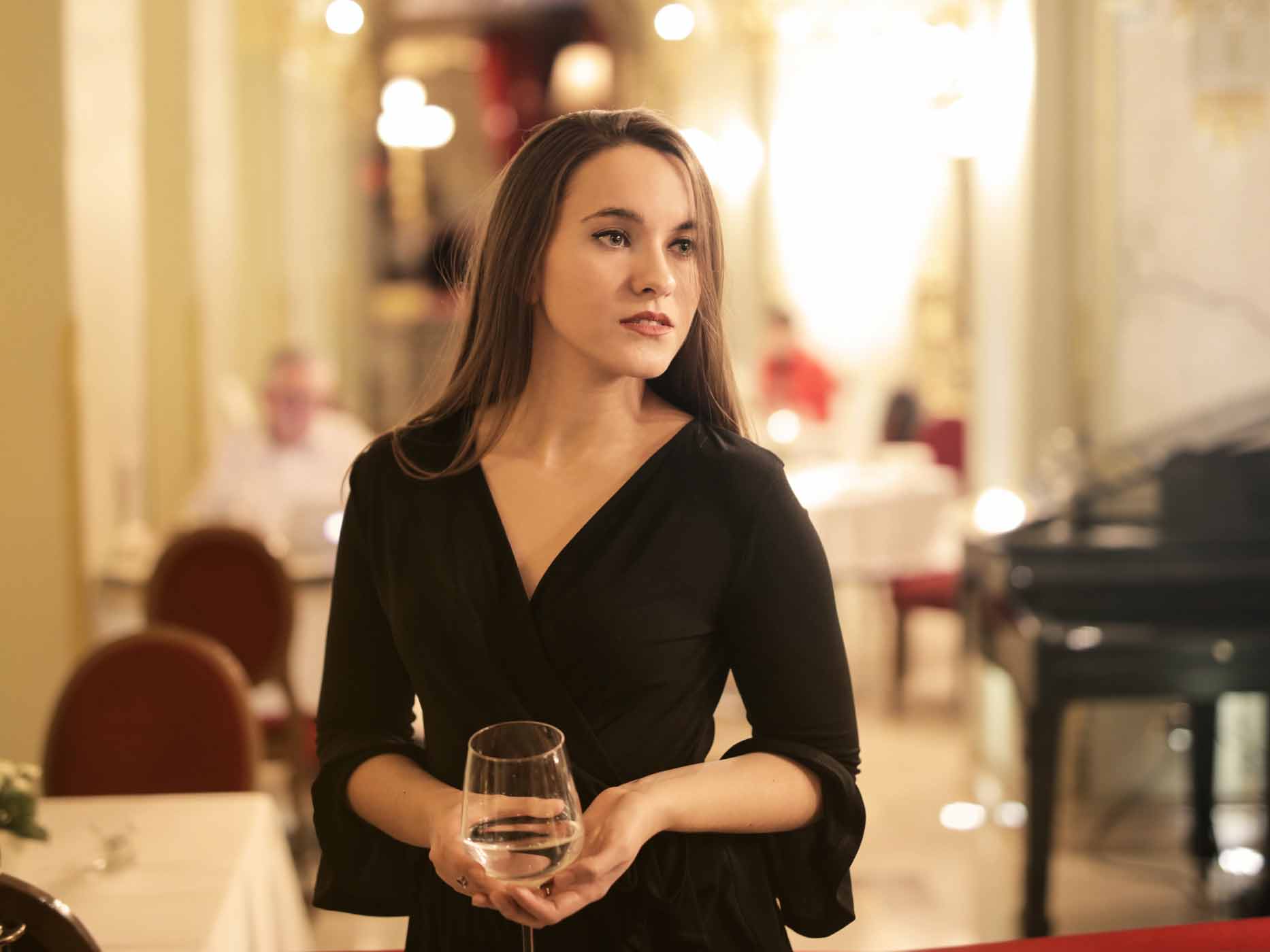Frau trägt elegantes, schwarzes Kleid in einem schicken Restaurant und hält dabei ein Glas Wein in der Hand