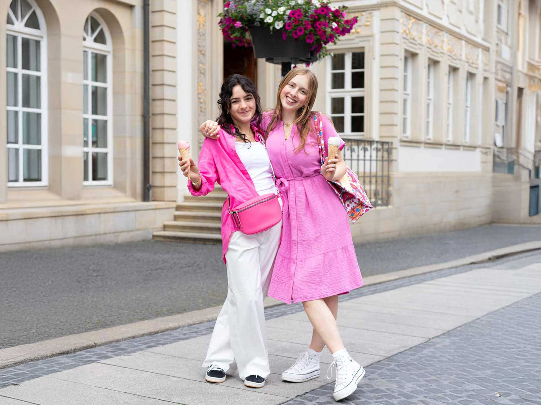 Zwei junge Mädchen tragen rosa und beige Kleidung. Die eine ein Kleid, die andere ein Top mit einer Hose. Sie essen jeweils ein Eis.