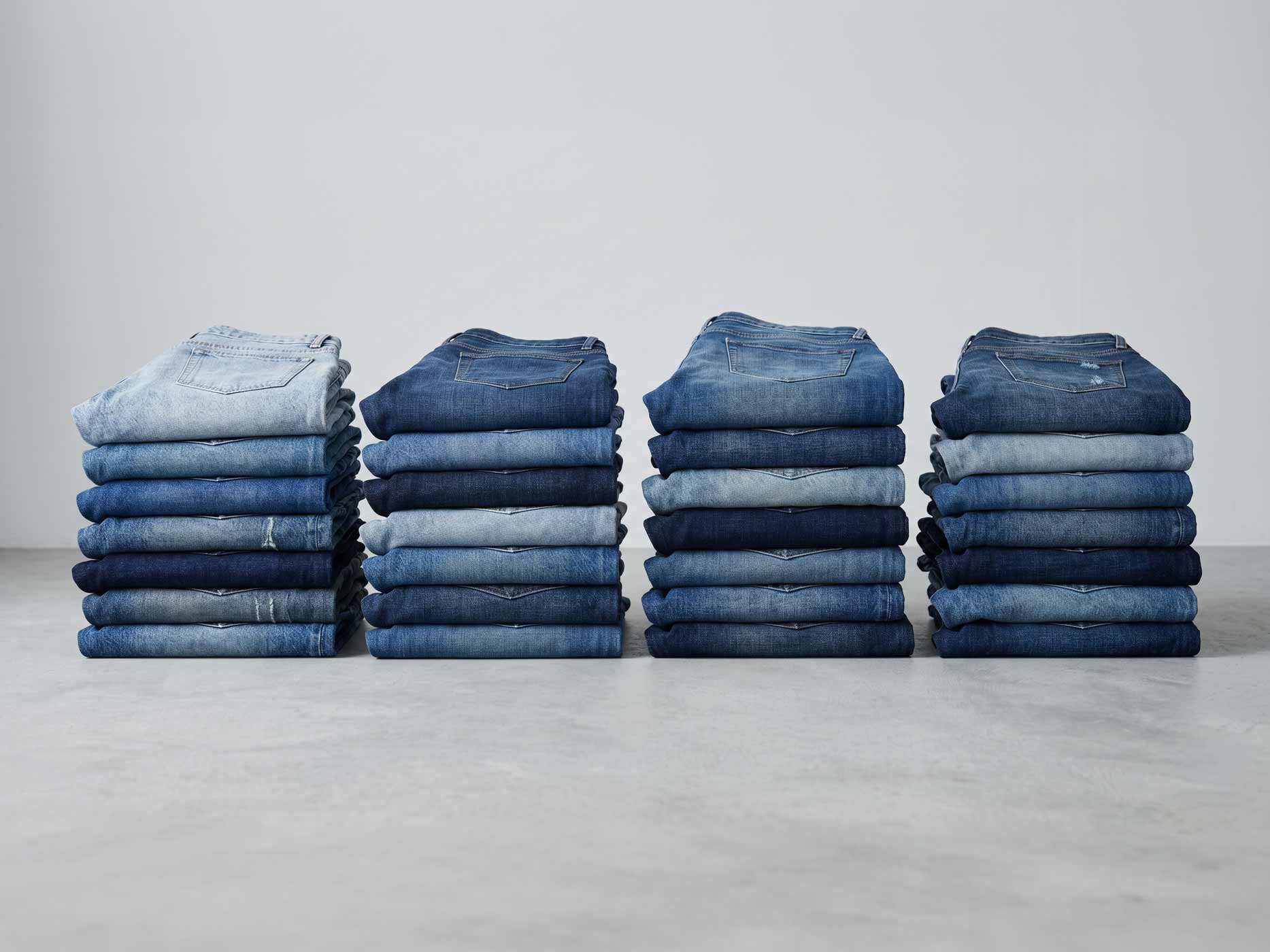 Mehrere Stapel gefalteter Jeans in unterschiedlichen Blaut&ouml;nen stehen nebeneinander auf einer hellen Fl&auml;che. Die Aufnahme zeigt verschiedene Waschungen und Farbnuancen von Denim.